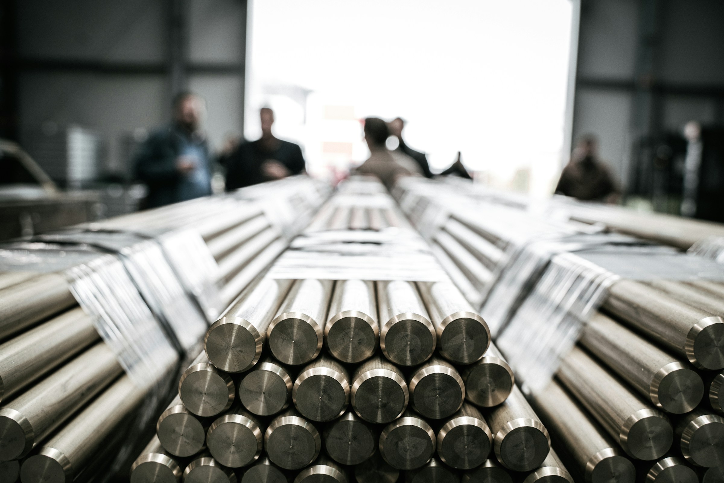 Stacks of cylindrical metal rods fill the foreground while several people stand in a workshop behind them.